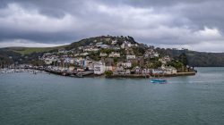 Dartmouth Harbour with boats and houses on a hill. The sea is in the foreground with dark skies.