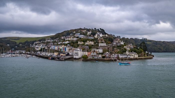 Dartmouth Harbour with boats and houses on a hill. The sea is in the foreground with dark skies.