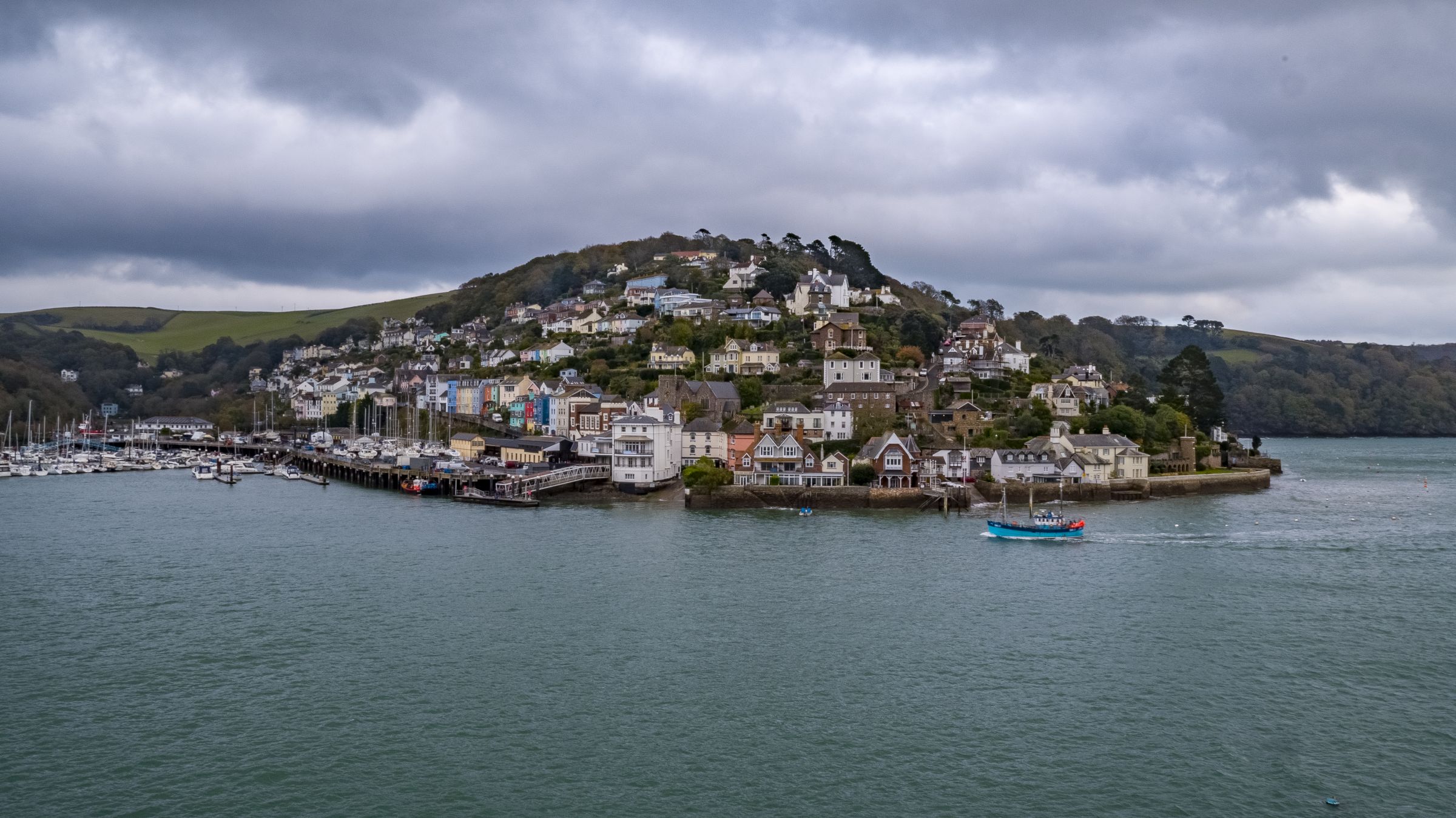 Dartmouth Harbour with boats and houses on a hill. The sea is in the foreground with dark skies.